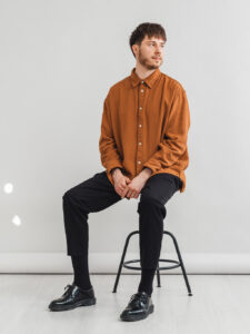Porträtt och CV Bilder i Linköping foto av man på stol i fotostudio. Orange skjorta och svarta byxor och skor tittar bort från kameran. Fotograferat av Satu Knape Fotograf i Linköping